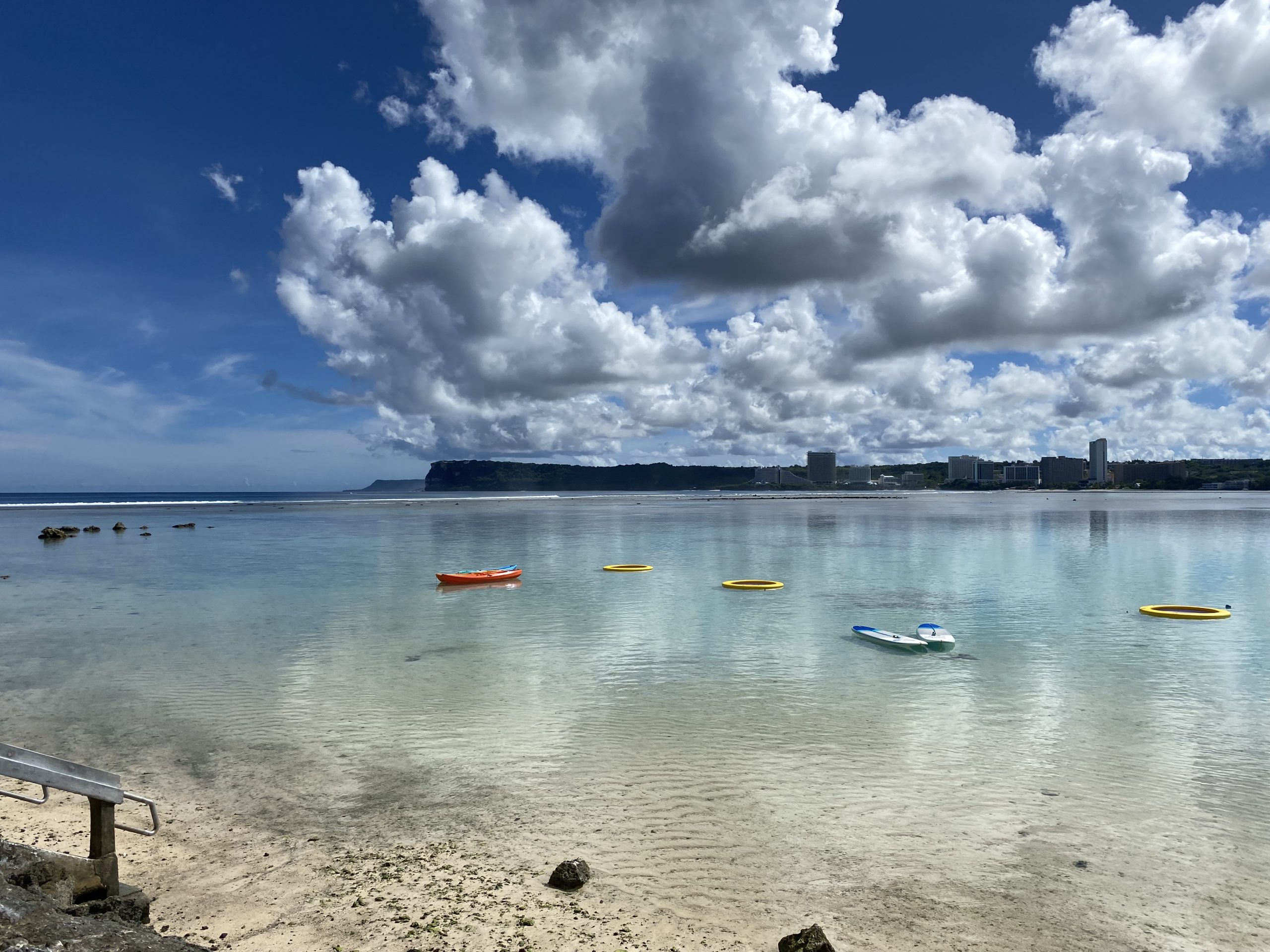 View of a beach with boats and tubes on the water and buildings in the distance.